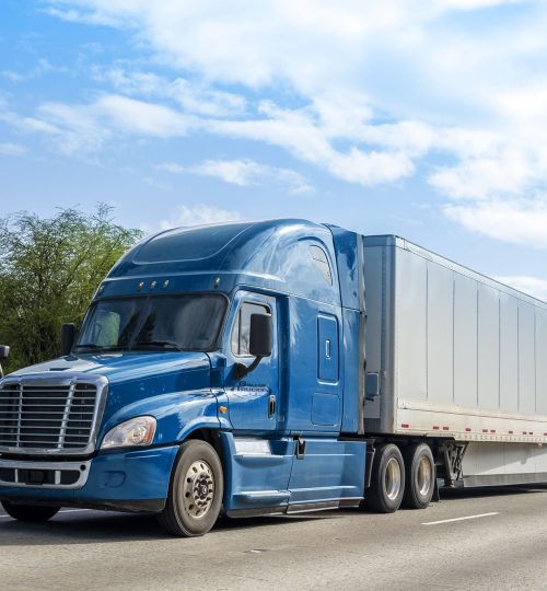 Red big rig popular professional reliable bonnet long haul semi truck transporting commercial cargo in dry van semi trailer moving on the straight wide highway with green trees on the background